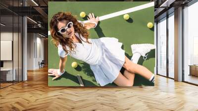 overhead view of fashionable tennis player in white sportswear and sunglasses resting on tennis court with balls and racket near by Wall mural