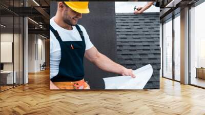 panoramic shot of handsome builder holding blueprint while coworker repairing roof Wall mural