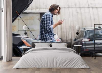 panoramic shot of man with smartphone standing near broken auto with open trunk, car insurance concept Wall mural