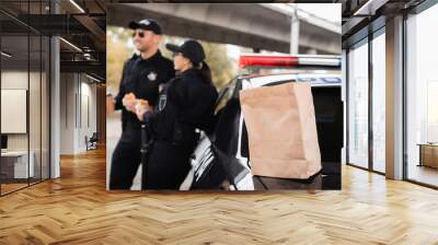 Paper bag on trunk of car near police officers with burgers and coffee to go on blurred background on urban street. Wall mural