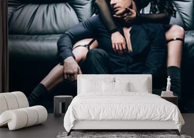 passionate man sitting on floor near sexy woman in bdsm costume on leather sofa isolated on black Wall mural