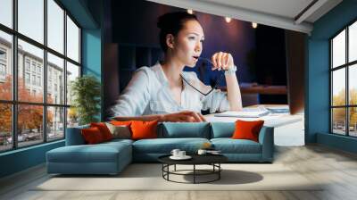 pensive asian businesswoman holding eyeglasses and looking at computer monitor, working late at the office Wall mural