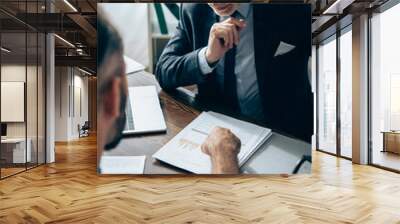 Pensive investor looking at papers with graphs near laptop and businessman on blurred foreground Wall mural