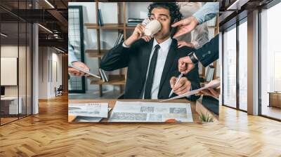 portrait of busy businessman drinking coffee and sitting at workplace while colleagues helping with  Wall mural