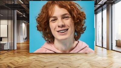 portrait of cheerful teenage guy with red wavy hair and braces smiling at camera on blue, happiness Wall mural