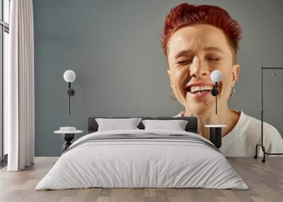 portrait of redhead bigender person with facial piercing laughing with closed eyes on grey backdrop Wall mural