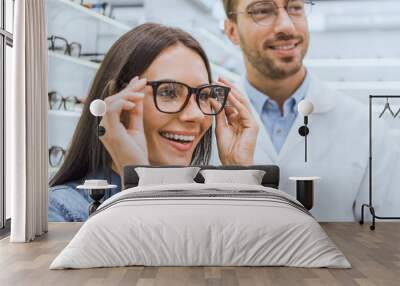 portrait of smiling woman choosing eyeglasses while male optometrist standing near in optics Wall mural