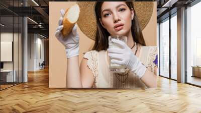 Portrait of stylish woman in sun hat holding milk and bread near butter isolated on beige Wall mural