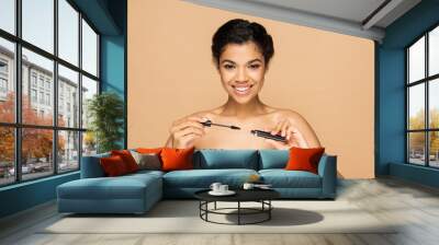 positive african american woman with bare shoulders holding mascara tube and brush isolated on beige Wall mural