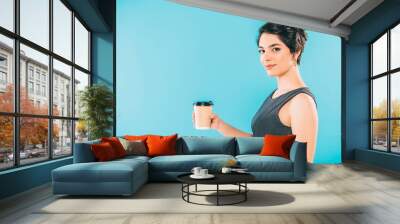 pretty mixed race woman holding paper cup while looking at camera on blue background Wall mural