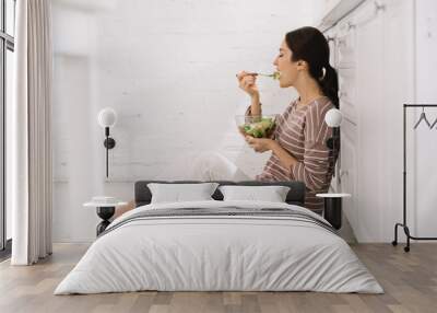 pretty young woman sitting on floor in kitchen and eating vegetable salad Wall mural