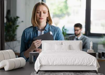 Sad woman holding paper folder with resume in blurred office. Wall mural