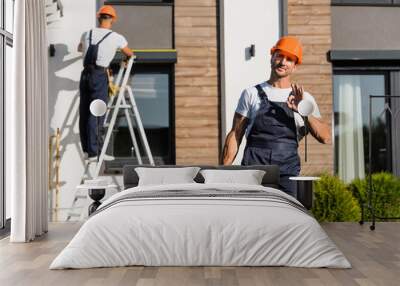 Selective focus of builder in uniform with toolbox showing ok gesture while colleague working near building Wall mural