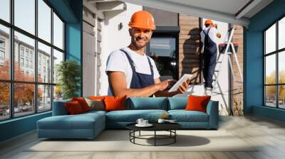 Selective focus of builder with digital tablet looking at camera while colleague working on ladder near house at background Wall mural