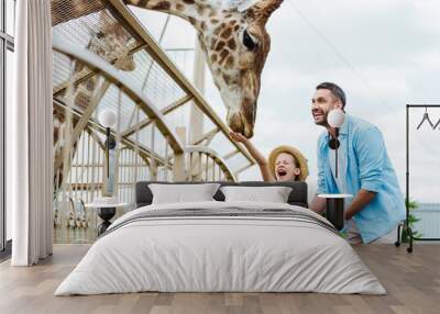 Selective focus of cheerful man and kid with closed eyes feeding giraffe in zoo Wall mural