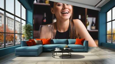 Selective focus of excited woman holding glass of wine while looking at camera at table in restaurant Wall mural