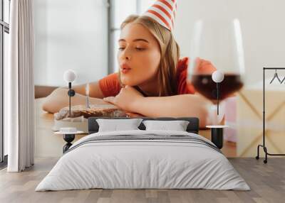 selective focus of girl in party cap blowing out candle on birthday cake near presents and wine glass on table Wall mural