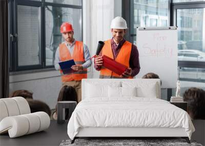 Selective focus of handsome firemen in helmets standing near white board with fire safety lettering and holding extinguisher and clipboard in hands Wall mural