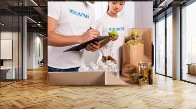 Selective focus of man writing on clipboard near asian volunteer holding apples and packages in charity center Wall mural