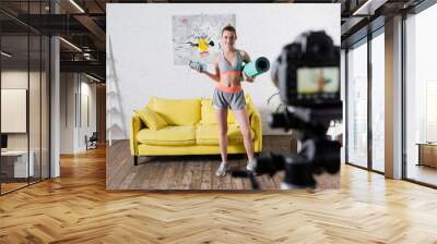 Selective focus of smiling sportswoman holding sports bottle and fitness mat near digital camera Wall mural