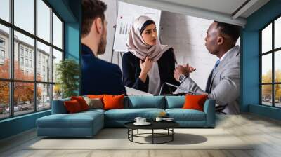 serious arabian businesswoman gesturing during meeting with african american business partner near translator, blurred foreground Wall mural