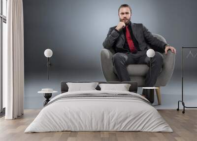 Serious young businessman sitting in armchair with hand to chin and looking to camera, isolated on gray Wall mural