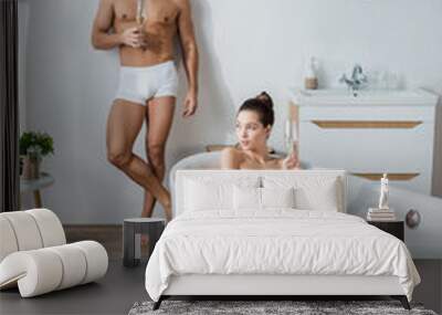 sexy man in underpants standing with champagne glass near girlfriend relaxing in bathtub. Wall mural