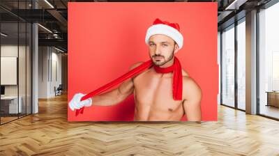 sexy muscular shirtless man in christmas hat and red scarf looking at camera isolated on red background Wall mural