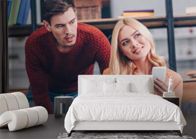 shocked husband looking at beautiful smiling wife using smartphone at home, jealousy concept Wall mural