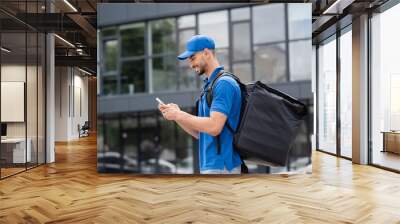 Side view of cheerful arabian courier with thermo backpack using smartphone on urban street Wall mural