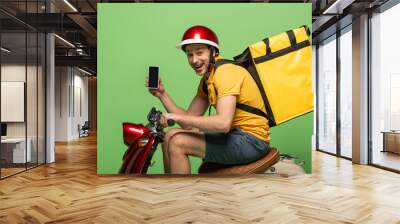 side view of happy delivery man in yellow uniform with backpack showing smartphone with blank screen on scooter isolated on green Wall mural