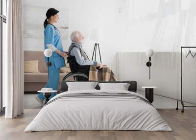 Side view of happy nurse standing in room with copy space, and carrying about disabled grey haired man in wheelchair Wall mural