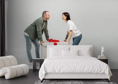 Side view of positive tattooed woman in casual clothes looking at husband while standing near big gift box during child protection day celebration on grey background Wall mural