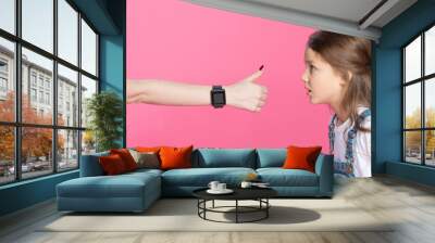 Side view of shocked little girl looking at woman with smartwatch showing thumb up Wall mural