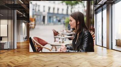 side view of smiling freelancer in black jacket looking at laptop near cup of coffee on table in french outdoor cafe. Wall mural