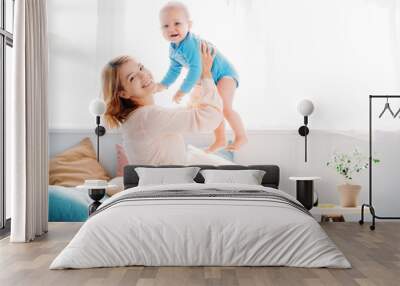 side view of smiling mother raising laughing little child while sitting on bed at home Wall mural