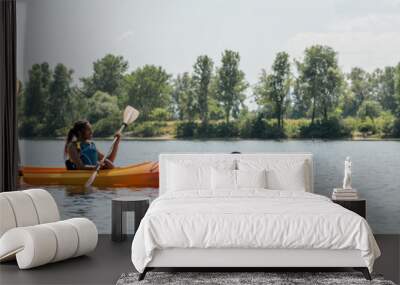 side view of young man and african american woman in life vests sailing in sportive kayak during active summer weekend on picturesque river with green trees on bank, banner Wall mural