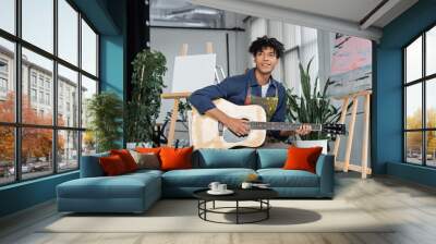 Smiling african american artist in dirty apron playing acoustic guitar near easels in studio. Wall mural