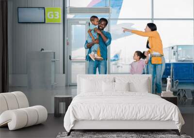 smiling african american family walking with baggage along waiting hall in airport while mother pointing with finger at son Wall mural