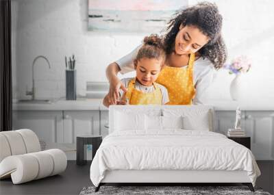 smiling african american mother helping daughter mixing eggs for dough in kitchen Wall mural