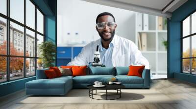 Smiling african american scientist in protective goggles looking at camera near microscope in lab Wall mural