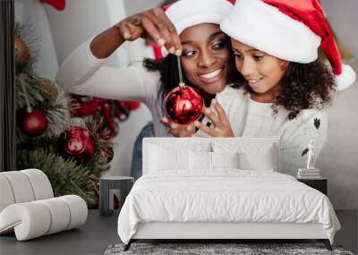 Smiling African American woman in Santa Claus hat helping daughter to decorate Christmas tree at home Wall mural