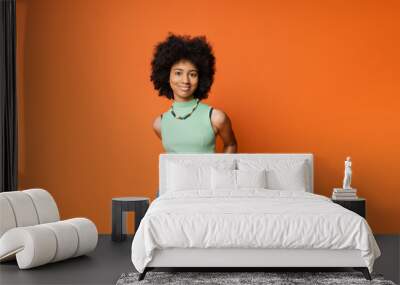 Smiling and stylish african american teenage girl in casual clothes with necklace and bold makeup posing and looking at camera isolated on orange, trendy teenage girl expressing individuality Wall mural