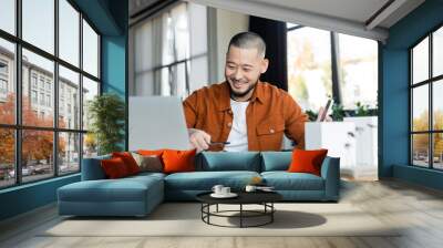 smiling asian architect holding smartphone while working with blueprints near blurred laptop Wall mural