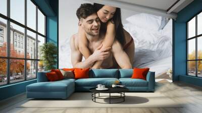 smiling asian woman hugging handsome shirtless boyfriend sitting on bed in blue jeans Wall mural