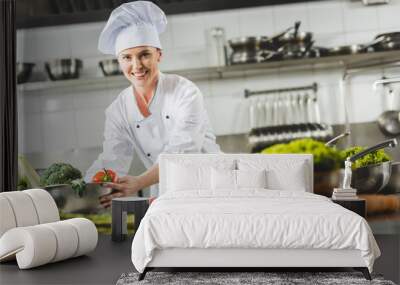 smiling attractive chef taking bowl with vegetables and looking at camera at restaurant kitchen Wall mural