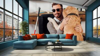 Smiling blind man sitting in armchair and hugging golden retriever at home Wall mural