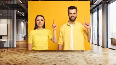 Smiling couple looking at camera while pointing with fingers isolated on yellow Wall mural