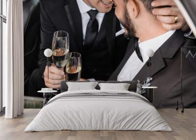 Smiling homosexual groom in formal wear holding champagne glass and hugging bearded boyfriend while celebrating wedding in car during honeymoon Wall mural