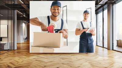 smiling indian mover packing box near coworker with digital tablet on blurred background Wall mural